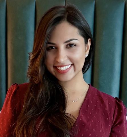 Smiling woman with long dark hair wearing a burgundy dress, seated in front of a green upholstered background.
